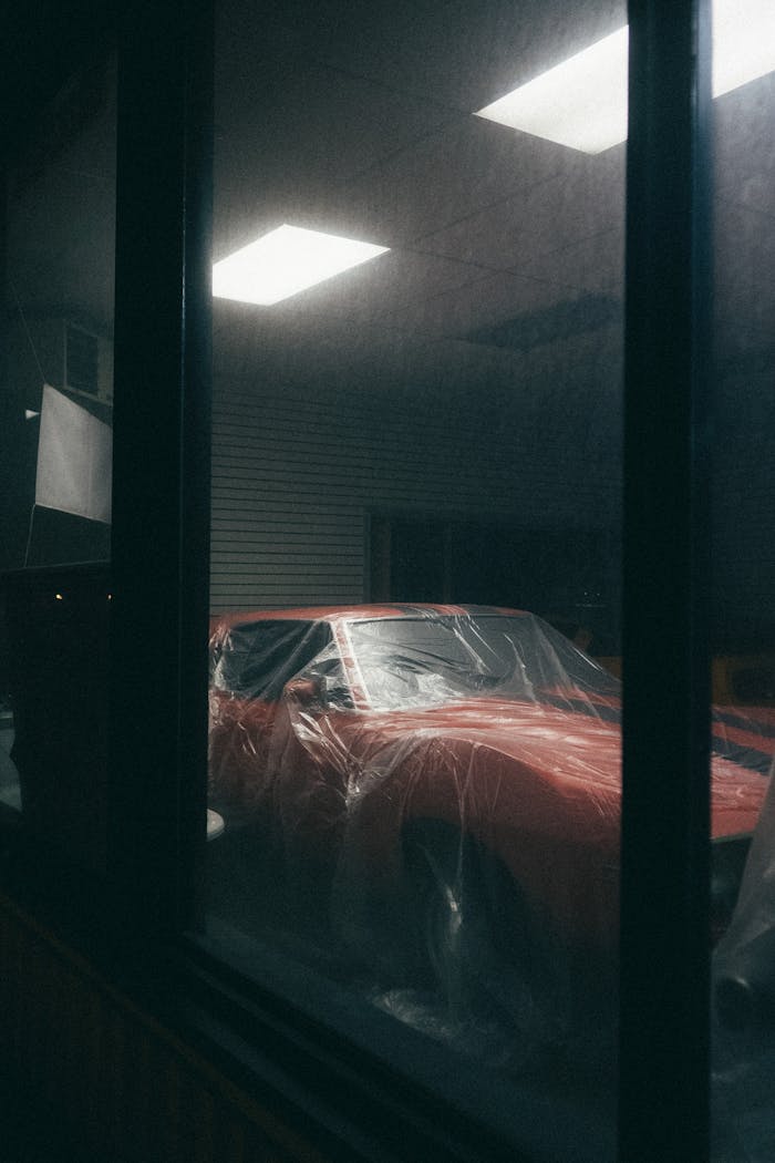 A dimly lit indoor scene showcasing a classic red car covered with protective plastic.